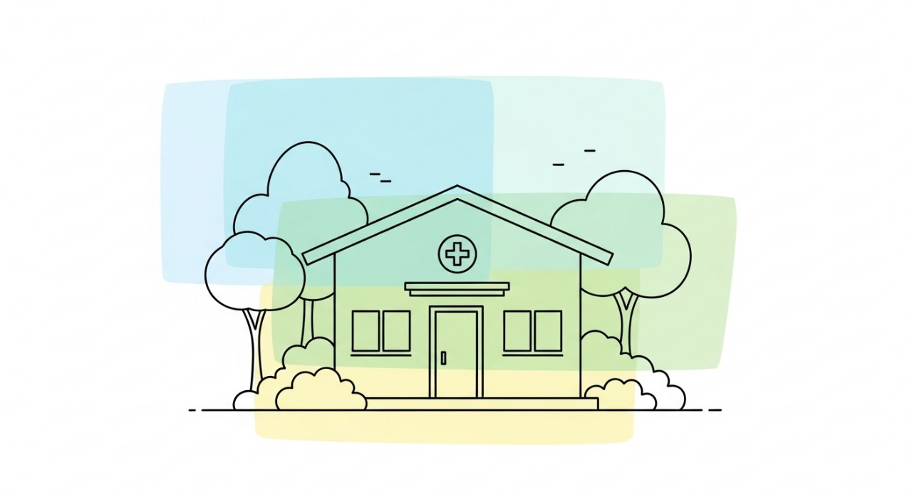 Pemandangan luar sebuah bangunan klinik desa yang kecil, bersih, dan dikelilingi sedikit kehijauan.